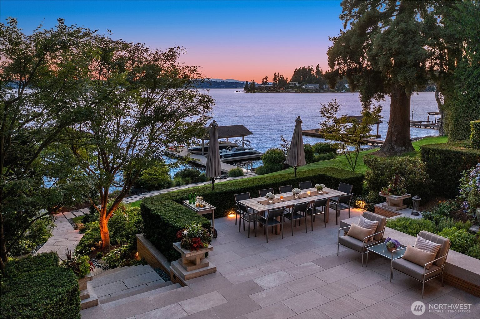 This stunning waterfront patio offers a sophisticated outdoor dining and lounge area, perfectly positioned to capture breathtaking sunset views over the lake. The space features a large stone-paved terrace, a formal dining set for ten, and comfortable lounge seating, all framed by manicured hedges and lush landscaping. The perspective is elevated, looking out from the terrace toward the private dock and the serene water beyond, creating an atmosphere of luxury and tranquility.