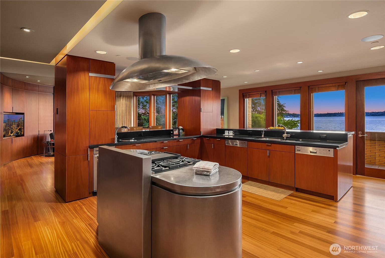 This sophisticated, modern kitchen features rich cherry wood cabinetry, sleek stainless steel appliances, and a prominent curved stainless steel island with a built-in cooktop. The space is defined by warm hardwood flooring and expansive windows that provide a stunning, panoramic view of the lake at sunset. The open-concept layout and high-end finishes create an inviting, luxurious atmosphere perfect for both cooking and entertaining.