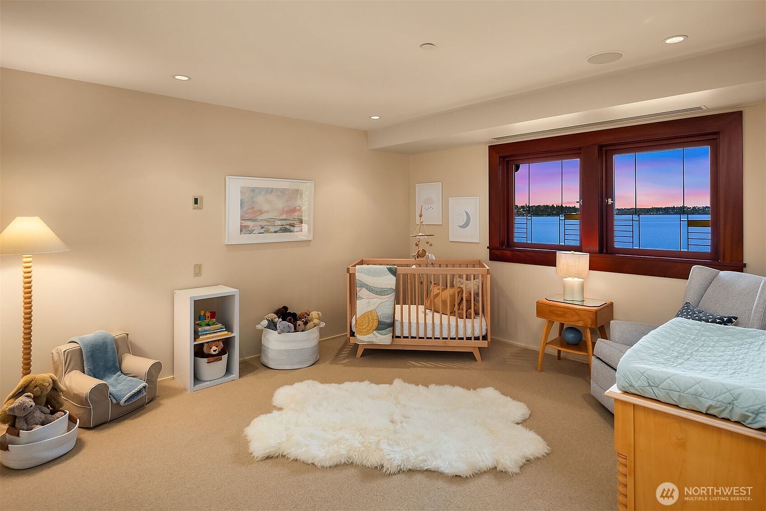 This cozy nursery features a light-toned wood crib as the focal point, complemented by a soft, plush white area rug and neutral-colored walls. A large window with dark wood trim provides a stunning, panoramic view of the water at sunset, adding a serene and peaceful atmosphere to the room. The space is thoughtfully furnished with a small armchair, a bookshelf, and a side table, creating a warm and inviting environment for a child.