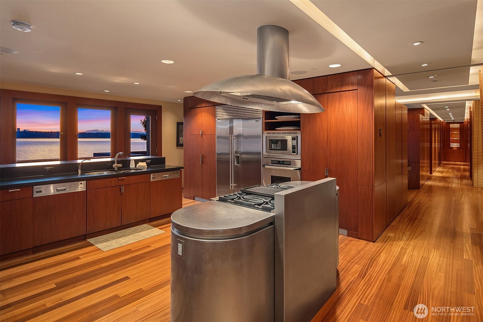 This sophisticated, modern kitchen features rich wood cabinetry and a prominent stainless steel island with an integrated gas cooktop and overhead range hood. The space is defined by its warm, polished hardwood floors and a seamless transition to a scenic waterfront view through large windows. The overall aesthetic is sleek and high-end, emphasizing clean lines and premium materials.