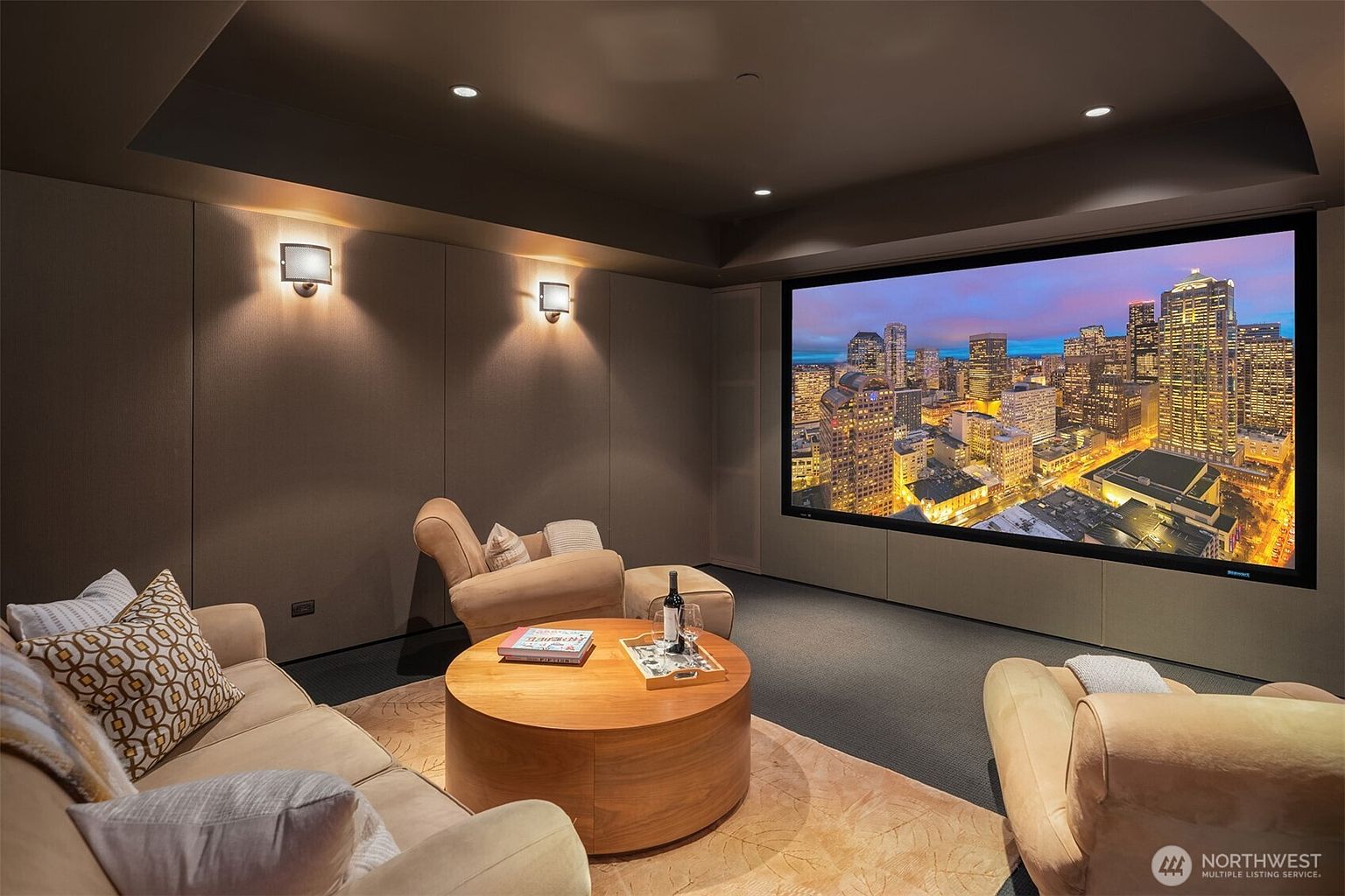 This sophisticated home theater room features a large, wall-mounted projection screen displaying a vibrant cityscape, creating an immersive cinematic experience. The space is furnished with plush, neutral-toned seating arranged around a circular wooden coffee table, set atop a textured area rug. With recessed ceiling lighting and warm wall sconces, the room offers a cozy, high-end atmosphere perfect for entertainment.