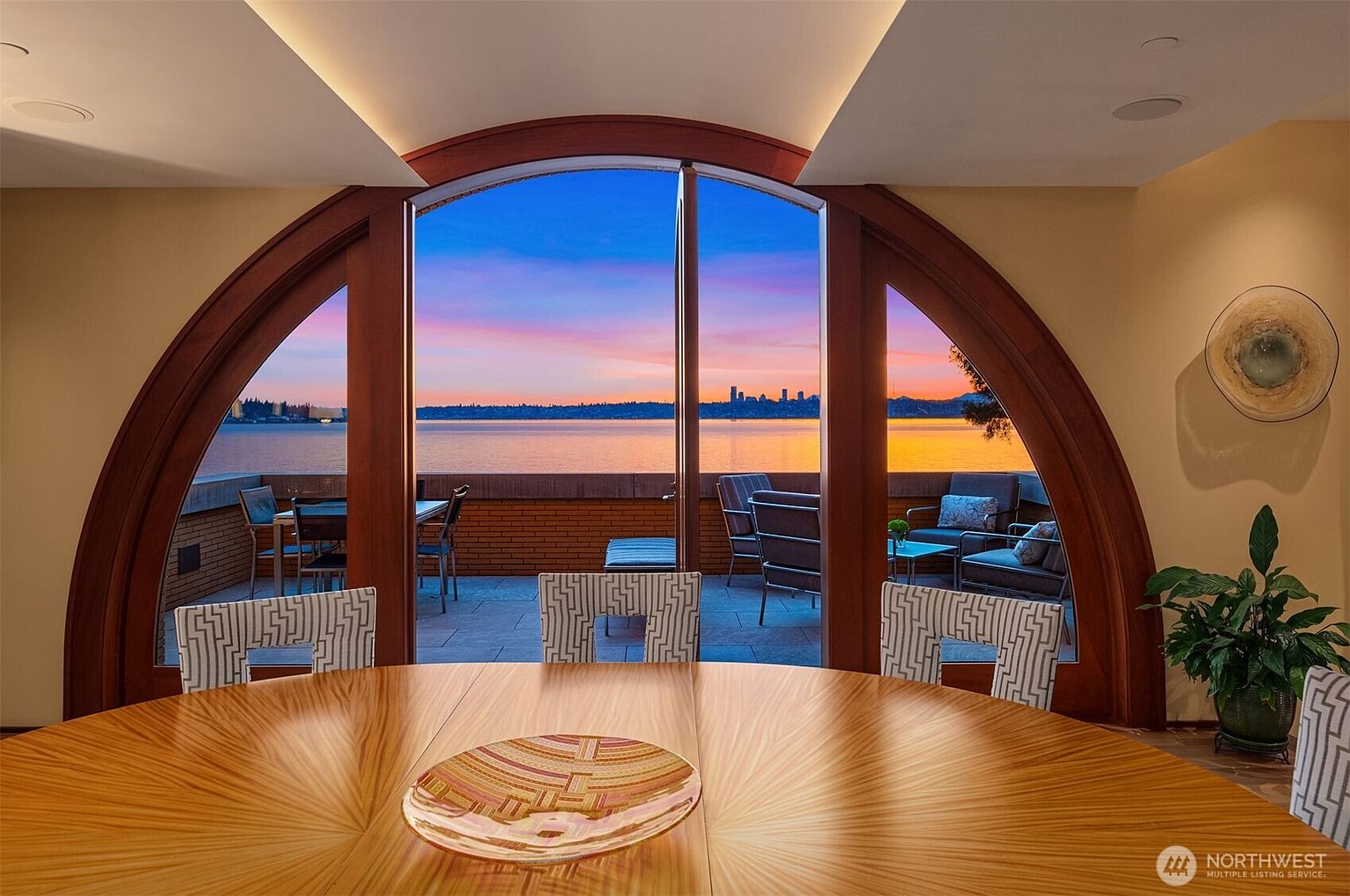 This elegant dining room features a striking, custom-designed circular wooden table with a unique grain pattern, centered in front of a massive, arched floor-to-ceiling window. The window frames a breathtaking sunset view over a calm body of water and a distant city skyline, seamlessly connecting the interior space to the outdoor patio. The room is characterized by its sophisticated, warm aesthetic and architectural grandeur, creating a serene and luxurious atmosphere.