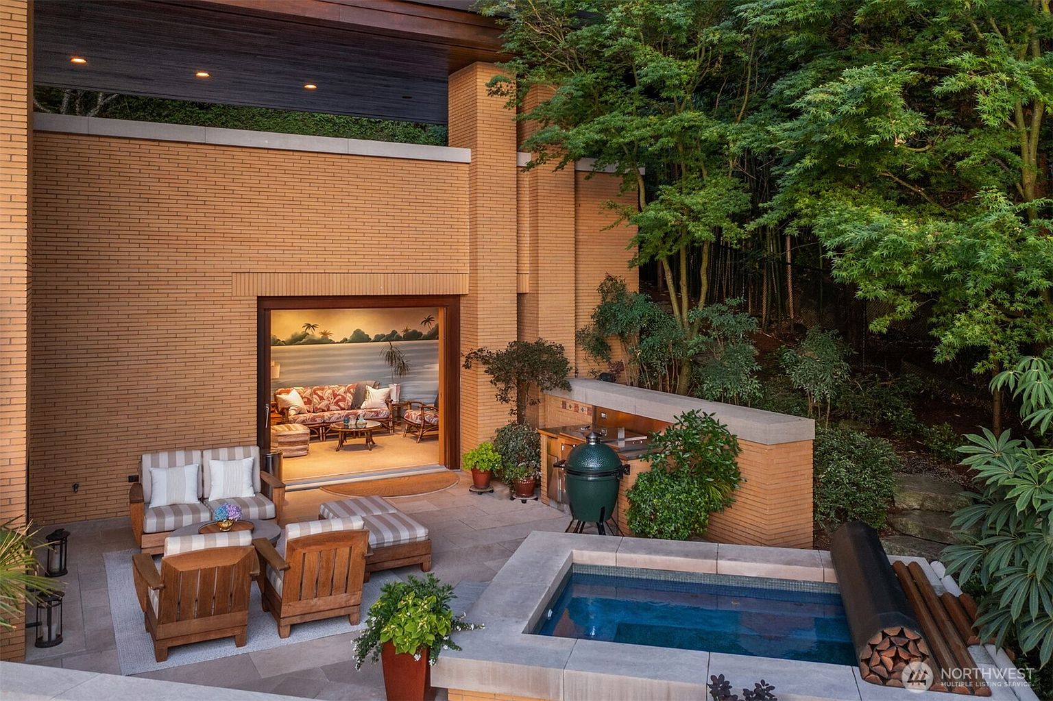 This sophisticated outdoor living space features a seamless transition from a cozy indoor lounge to a spacious brick-paved patio. The area is well-appointed with comfortable striped outdoor furniture, a built-in grilling station with a green ceramic kamado-style grill, and a sleek, stone-rimmed spa. Surrounded by lush greenery and mature trees, the setting offers a private, tranquil retreat perfect for relaxation and entertaining.