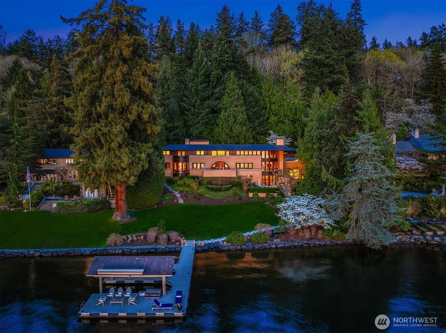 This stunning aerial view captures a luxurious waterfront estate at twilight, nestled against a dense, lush forest backdrop. The multi-level residence features warm, inviting interior lighting that contrasts beautifully with the cool blue tones of the lake and evening sky. A private dock with a covered seating area extends into the water, while a manicured lawn and mature trees frame the property, creating an atmosphere of serene, high-end seclusion.