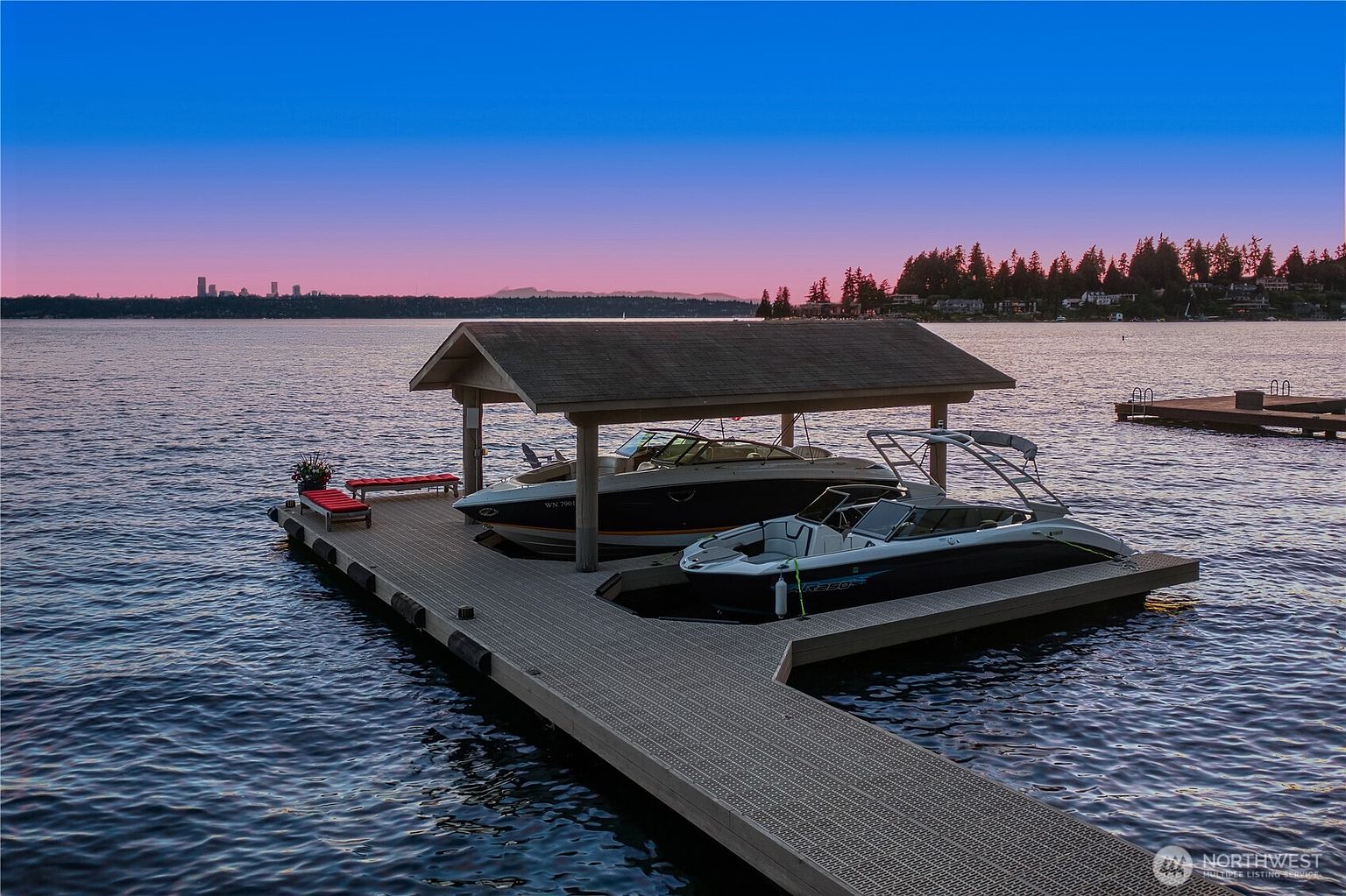 This aerial perspective captures a private boat dock on a calm lake at sunset, featuring a covered slip housing two modern motorboats. The scene is framed by a vibrant, gradient sky transitioning from deep blue to soft pink, with a distant city skyline visible on the horizon. The composition emphasizes a serene, luxurious waterfront lifestyle, highlighting the well-maintained dock structure and the peaceful, expansive water.