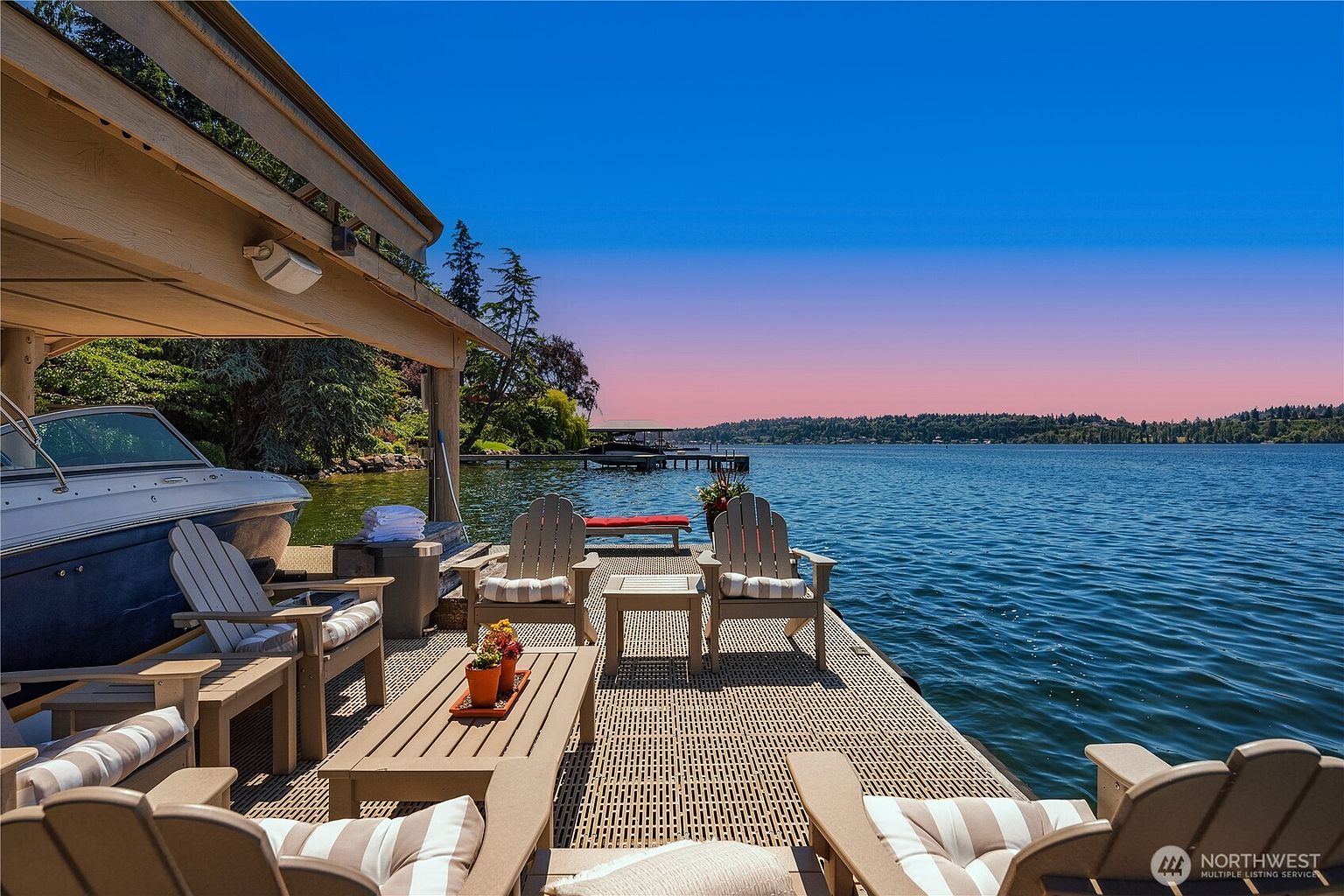 This serene waterfront deck features a covered boat slip and a comfortable seating area furnished with Adirondack chairs and a wooden coffee table. The perspective captures a tranquil, expansive view of the lake under a vibrant sunset sky, highlighting the property's prime location for relaxation and recreation. The overall atmosphere is peaceful and luxurious, emphasizing the seamless connection between the home's outdoor living space and the water.