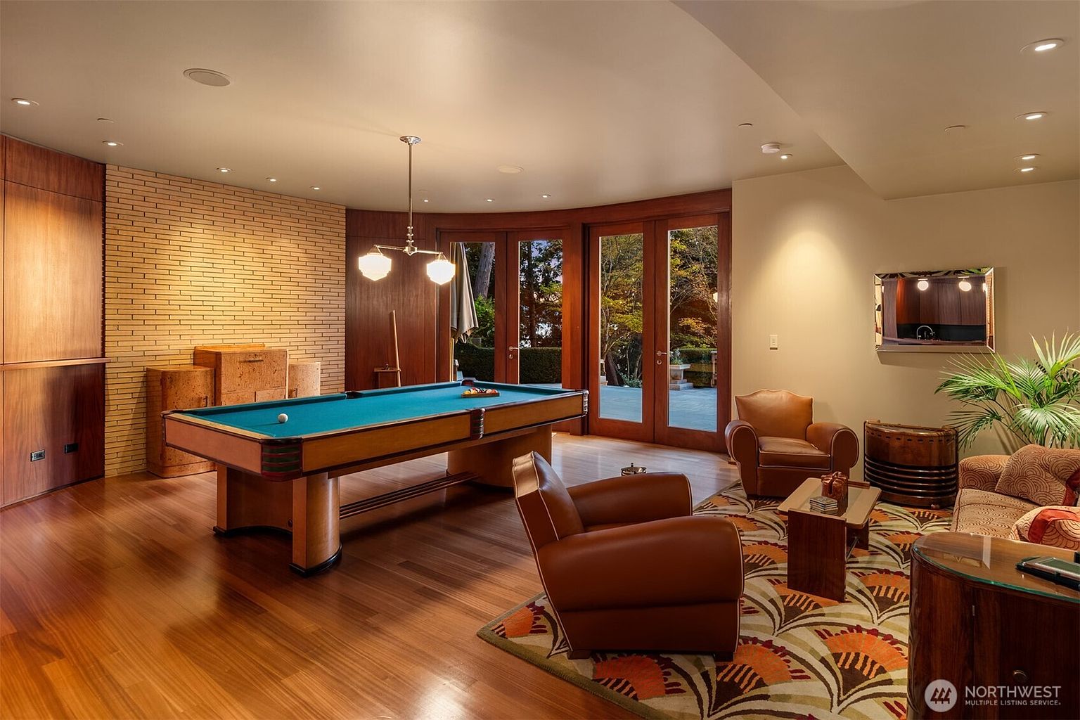 This sophisticated game room features a prominent pool table as its centerpiece, set against a backdrop of warm wood paneling and a textured brick accent wall. The space is furnished with comfortable leather armchairs and a patterned area rug, creating a cozy yet upscale atmosphere that flows seamlessly to an outdoor patio through large glass doors. The room's warm lighting and rich materials evoke a classic, refined aesthetic perfect for entertainment.