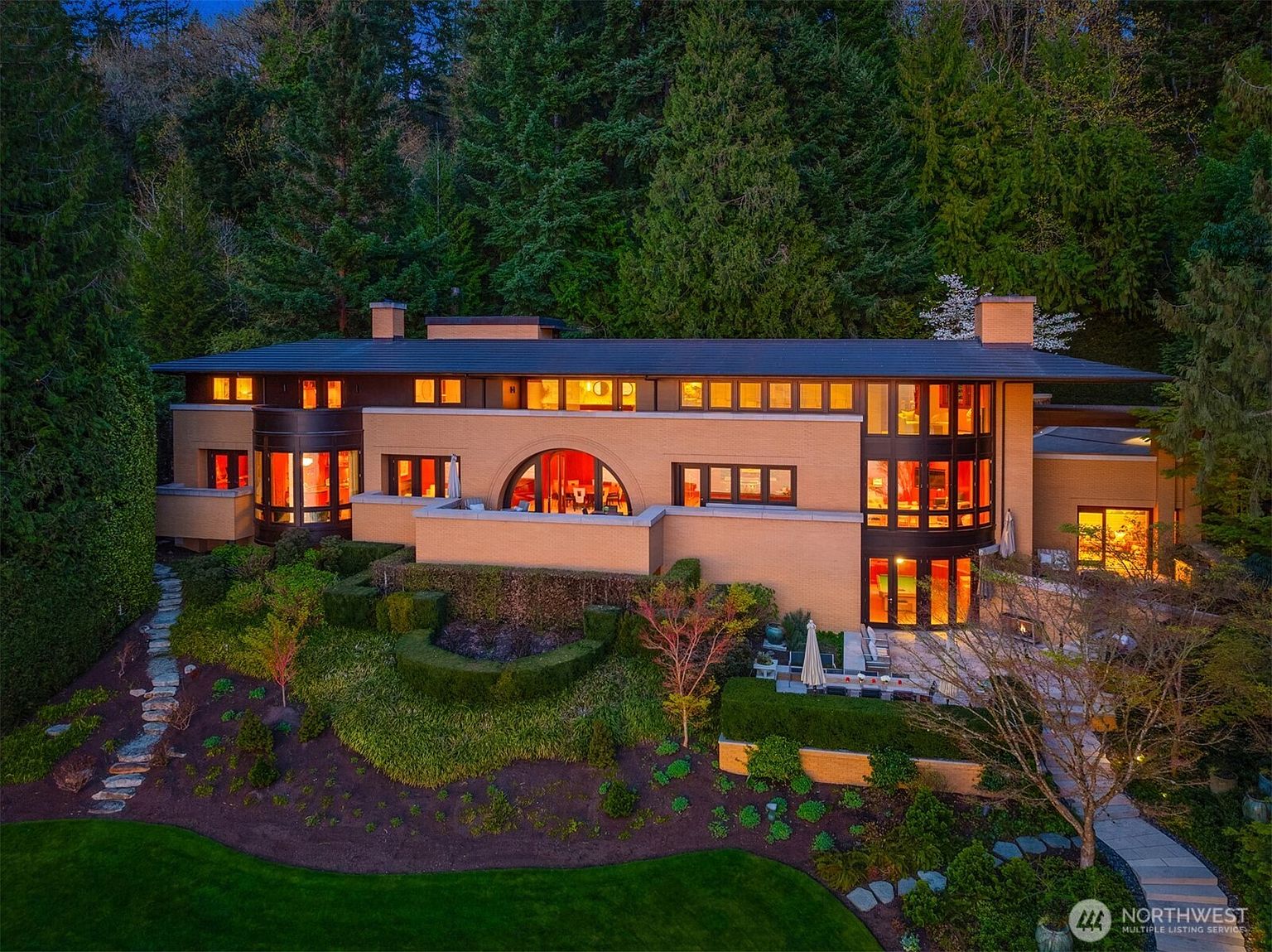 This stunning architectural residence showcases a sophisticated blend of Prairie-style design with modern luxury, featuring expansive glass windows and warm brick masonry. The home is nestled against a lush, dense forest backdrop, with a beautifully landscaped terraced garden and a spacious stone patio area in the foreground. The warm interior lighting glowing through the windows creates an inviting and elegant atmosphere at dusk.