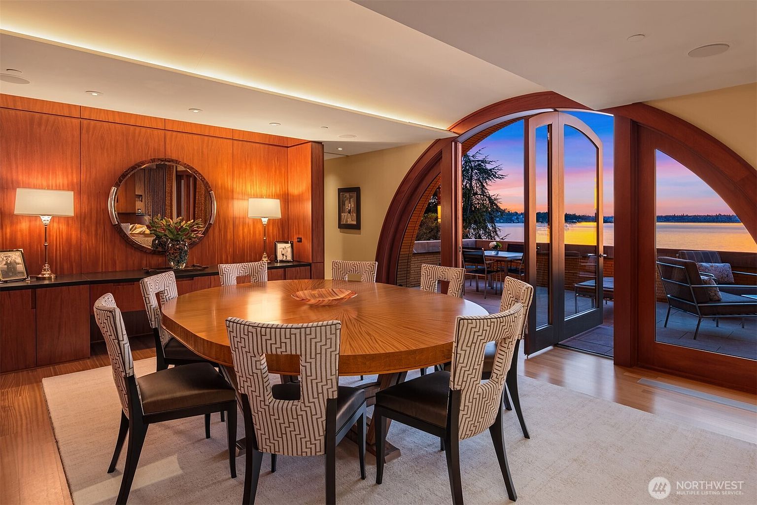 This elegant dining room features a large, circular wooden table surrounded by patterned chairs, set against a backdrop of rich wood-paneled walls and a circular mirror. A dramatic arched glass door opens onto a balcony, framing a breathtaking sunset view over the water. The space exudes a sophisticated, warm atmosphere, blending high-end craftsmanship with a seamless connection to the outdoor scenery.