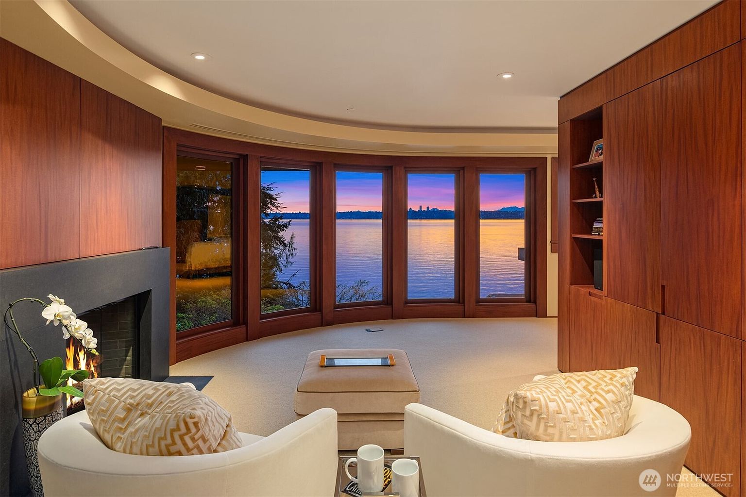 This sophisticated living room features a curved wall of floor-to-ceiling windows that frame a breathtaking sunset view over the water. The space is anchored by a sleek, modern fireplace and warm wood-paneled walls, complemented by plush, neutral-toned seating. The overall aesthetic is one of refined luxury and tranquility, perfectly designed to showcase the panoramic waterfront scenery.