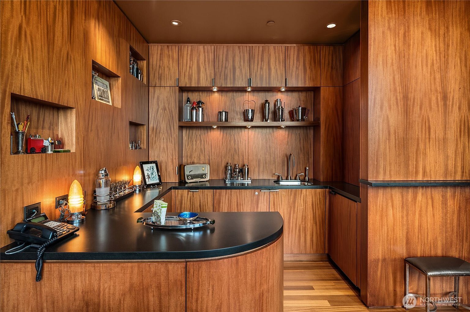 This sophisticated home office or wet bar area features rich, warm-toned wood paneling throughout, creating a luxurious and cohesive aesthetic. The space includes a curved black countertop, a built-in sink, open shelving for display, and recessed lighting that highlights the wood grain. A desk area with a telephone and lamp suggests a functional workspace, while the overall design exudes a refined, mid-century modern influence.