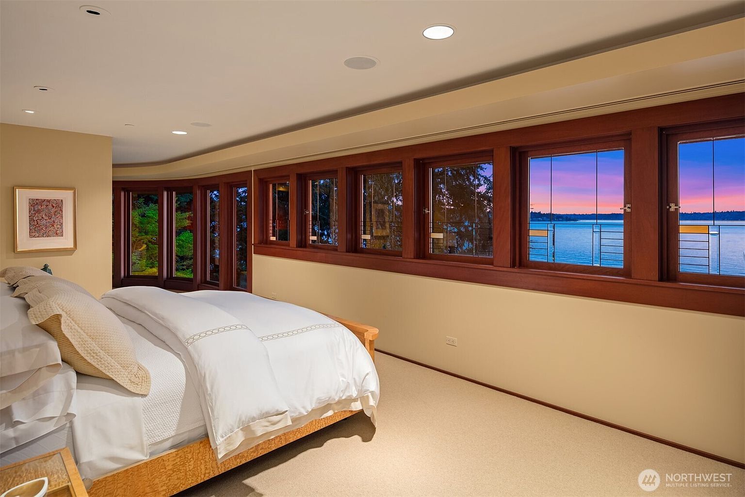 This primary bedroom features a plush, neutral-toned bed set against a wall of expansive, wood-framed windows that offer a stunning panoramic view of the water at sunset. The room is characterized by its warm, inviting atmosphere, soft carpeting, and recessed lighting, creating a serene and luxurious retreat. The perspective is from the corner of the room, emphasizing the seamless blend of comfortable indoor living and the scenic outdoor landscape.
