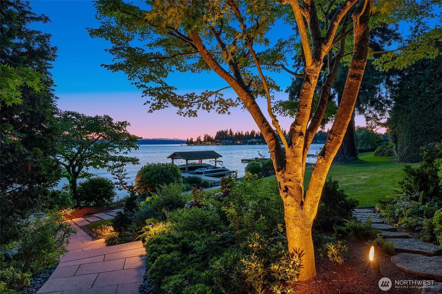 This stunning waterfront property features a beautifully landscaped garden with stone pathways leading toward a private dock on a serene lake at dusk. The scene is illuminated by warm, ambient landscape lighting that highlights the mature trees and lush greenery, creating a tranquil and inviting atmosphere. The perspective captures the seamless transition from the manicured lawn to the water's edge, emphasizing the home's premium lakeside location.