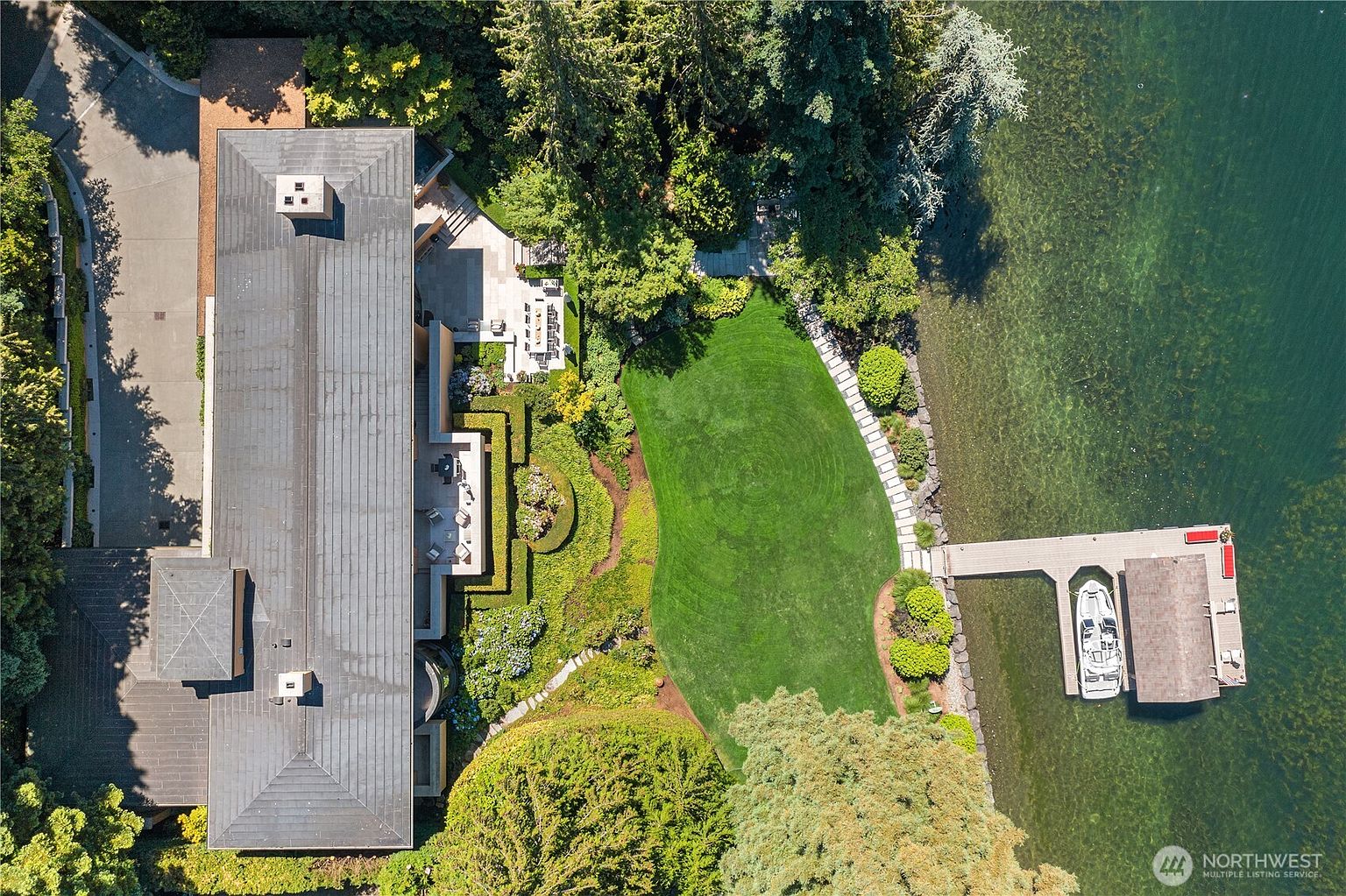 This high-angle aerial view captures a luxurious waterfront property, showcasing a long, modern residence with a dark roof nestled among lush, mature trees. The property features a meticulously landscaped lawn that curves toward the water's edge, leading to a private dock with a boat moored alongside. The perspective emphasizes the seamless integration of the home with its natural surroundings and the expansive, serene lakefront setting.