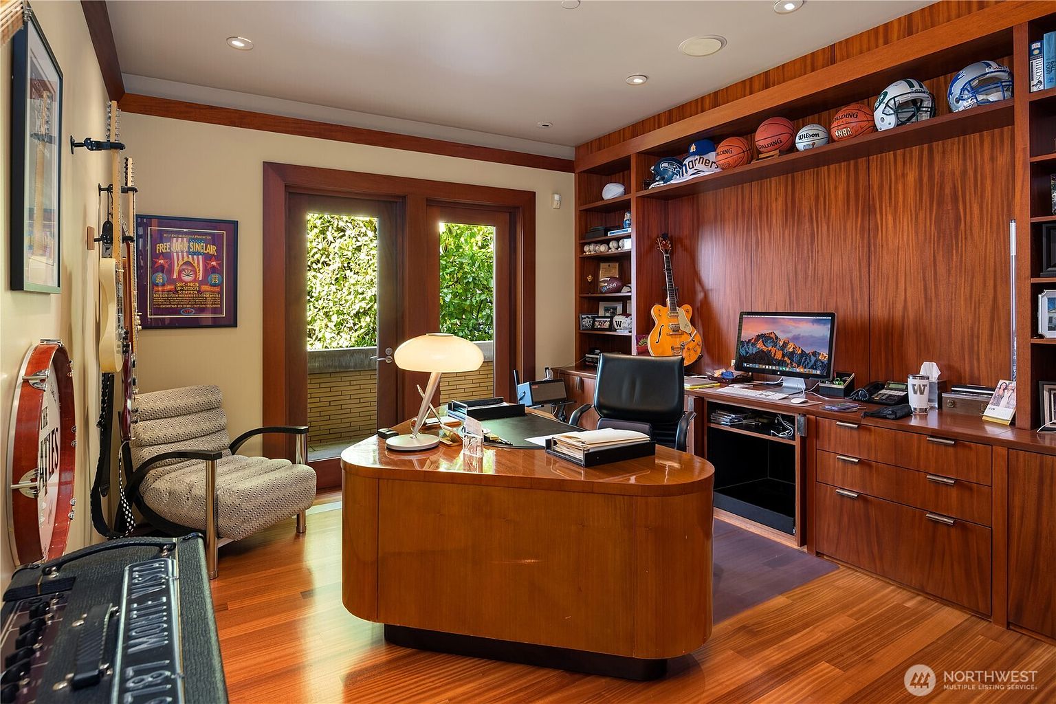 This sophisticated home office features rich, warm wood paneling and cabinetry that creates a professional and inviting atmosphere. The room is anchored by a large, custom-shaped wooden desk and includes built-in shelving displaying sports memorabilia and a guitar, alongside a comfortable armchair and a glass door leading to an outdoor space. The perspective is a wide, eye-level shot that captures the entire workspace, emphasizing the high-end finishes and personalized decor.