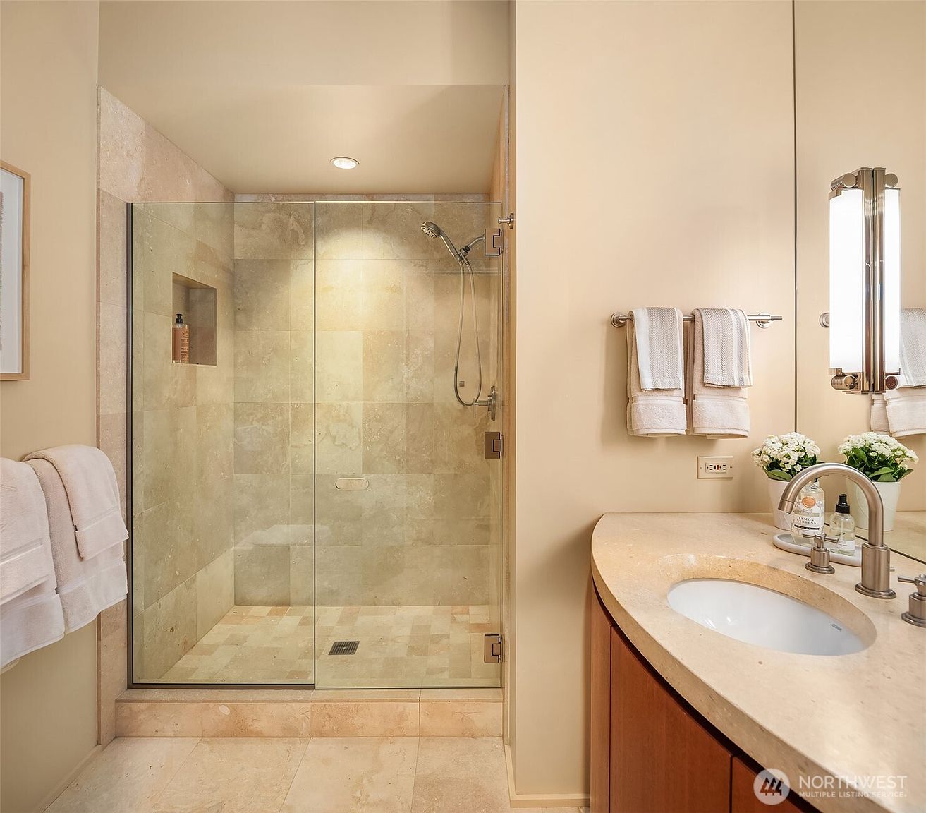 This elegant bathroom features a walk-in glass-enclosed shower with neutral-toned stone tile and a built-in niche. Adjacent to the shower is a vanity with a curved stone countertop, a modern faucet, and a large mirror, creating a clean and sophisticated aesthetic. The warm, neutral color palette and high-quality finishes provide a spa-like atmosphere.