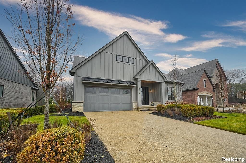 This is a front view of a modern, gray-toned house with a two-car garage and a well-manicured lawn. The house features a combination of vertical siding and stone accents, with a dark-colored roof and trim. The driveway leads directly to the garage, and there are neatly trimmed bushes and landscaping elements that enhance the curb appeal.