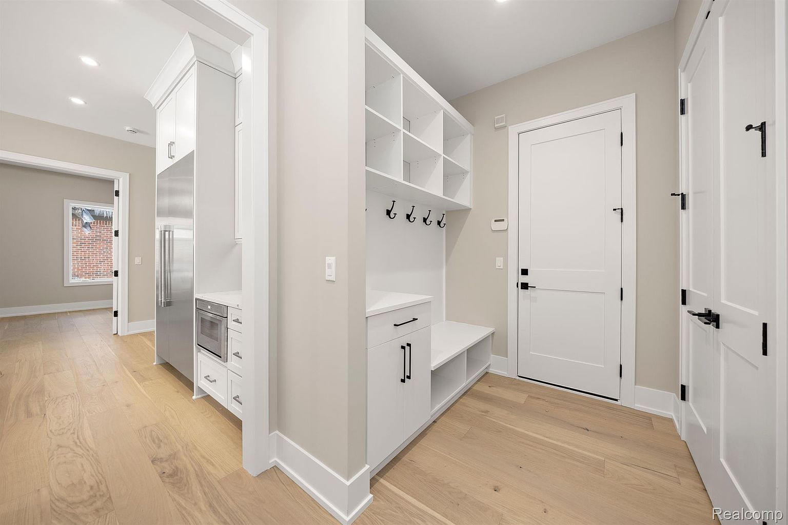 This interior shot showcases a well-lit hallway with light wood flooring and neutral-toned walls. Built-in storage solutions, including shelving and a bench with hooks, provide practical organization. A stainless steel refrigerator and microwave are integrated into the cabinetry, adding a modern touch to the space.