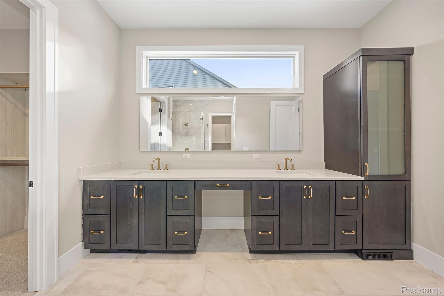 This is a well-lit primary bathroom featuring a double vanity with dark gray cabinetry and gold hardware. A large mirror spans the length of the vanity, reflecting a view into another room. The countertops are light-colored, and the flooring is a light marble tile, creating a clean and luxurious feel.