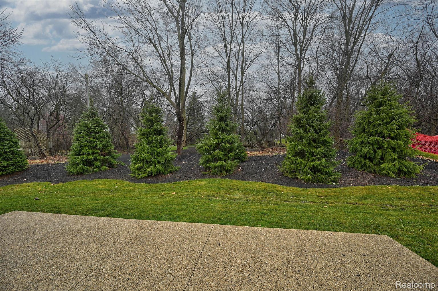 The image showcases a well-maintained yard with a concrete patio in the foreground, leading to a lush green lawn. A row of evergreen trees lines the back, set against a backdrop of bare trees and a cloudy sky. The scene presents a serene and inviting outdoor space, perfect for relaxation and entertainment.