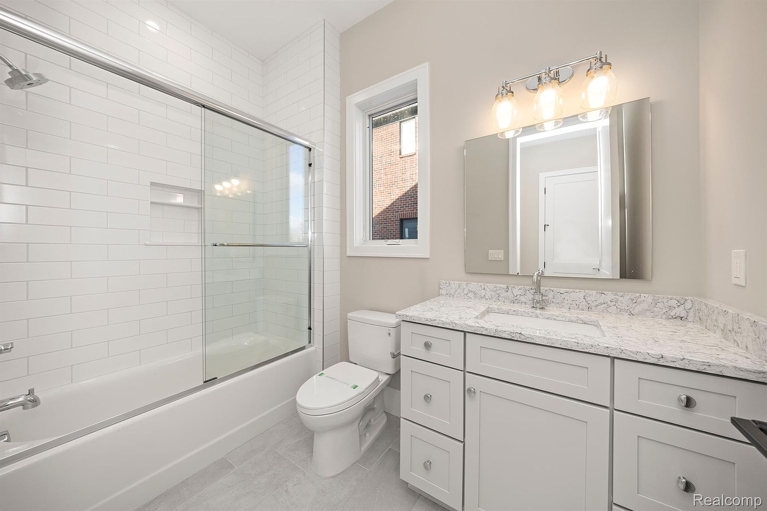 This is a well-lit bathroom featuring a bathtub with a glass shower door, a white toilet, and a vanity with a white countertop and white cabinets. The walls are painted in a neutral tone, and the floor is tiled. The overall impression is clean and modern, suitable for a guest or secondary bathroom.