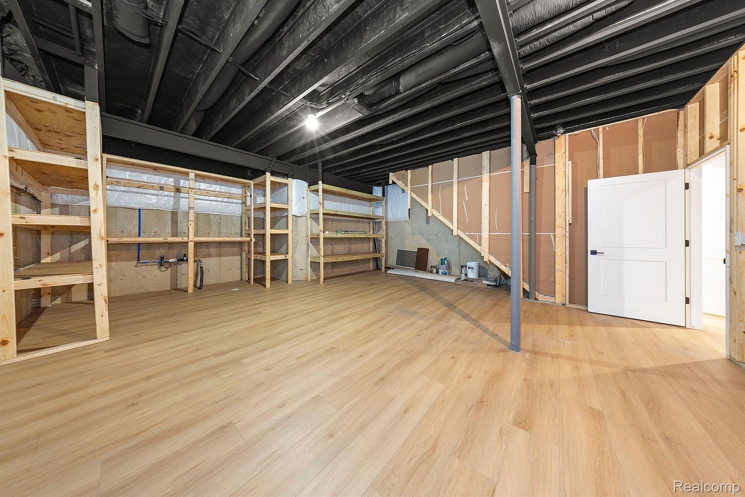 This is an interior shot of a basement. The basement features unfinished walls, exposed ceiling beams painted black, and light wood-look flooring. There are wooden shelves along the walls, and a white door is visible on the right side of the frame. The perspective is a wide shot, showcasing the spaciousness of the basement.