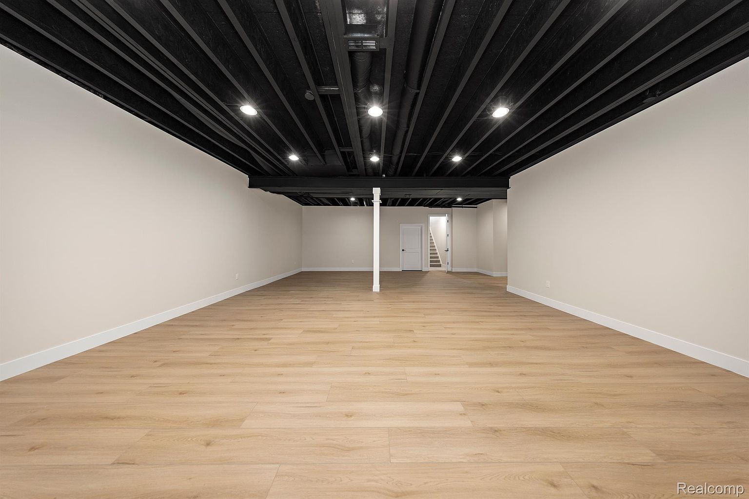 This is a spacious, finished basement with light wood-look flooring and neutral-colored walls. The ceiling is exposed and painted black, with recessed lighting throughout. A support beam is visible in the center of the room, and a doorway leads to a staircase, suggesting potential for additional living space or storage.