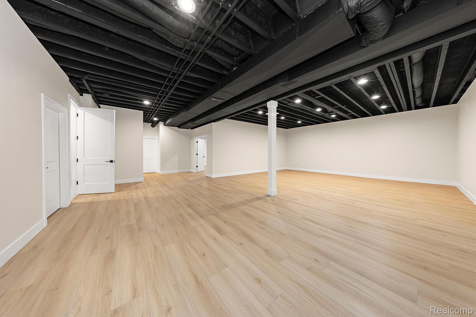 This is a spacious, newly renovated basement featuring light wood-look flooring and a black-painted exposed ceiling with recessed lighting. The walls are painted in a neutral tone, and there are several white doors leading to other rooms. A support beam is visible in the center of the room, and the overall impression is clean and modern.
