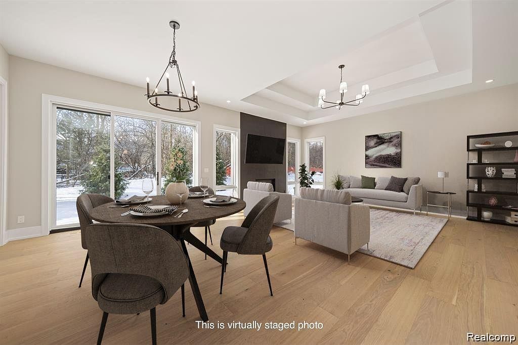 This is an interior shot of a living room and dining area. The dining area features a round table with modern chairs and a chandelier, while the living room has a gray sofa, armchairs, and a fireplace with a mounted TV. Large windows provide natural light and views of the outdoors, and the hardwood floors add warmth to the space.