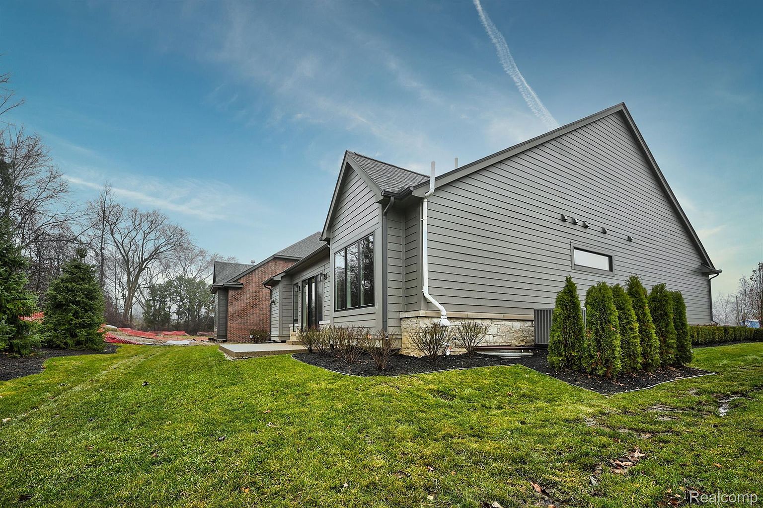 This is a side view of a modern house with gray siding and a dark gray roof. The house has a well-maintained lawn and landscaping, including small bushes and trees. The exterior features a combination of siding and stone accents, adding visual interest and texture.