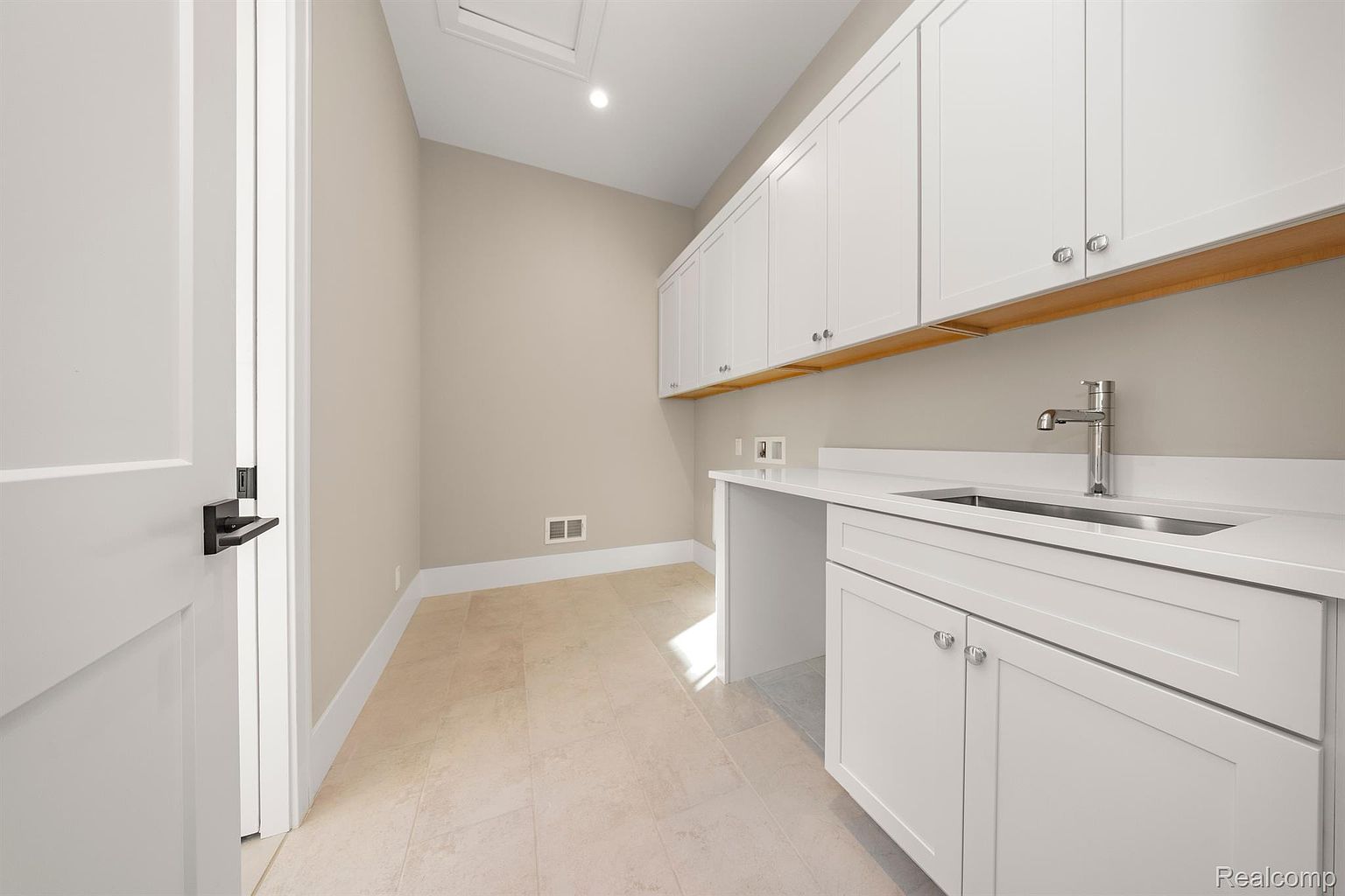 This is an interior shot of a laundry room. The room features white cabinets, a sink with a modern faucet, and light-colored tile flooring. The walls are painted in a neutral tone, and there is a door on the left side of the frame. The overall impression is clean and functional.