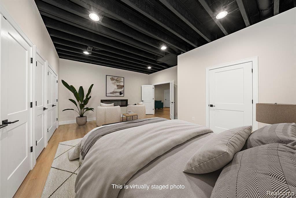 This is an interior shot of a bedroom featuring a bed with neutral-toned bedding, a black-painted exposed ceiling, and light-colored walls. There are several white doors with black hardware along one wall, and a glimpse into an adjacent room reveals a seating area with a fireplace. The room has a modern and minimalist aesthetic.