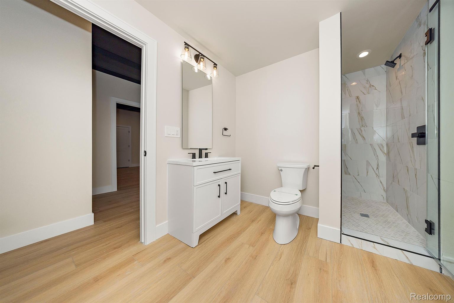 This is a well-lit bathroom featuring a modern design. The bathroom includes a white vanity with a dark countertop, a toilet, and a glass-enclosed shower with marble-patterned tiles. The flooring is a light wood-look tile, and the walls are painted in a neutral tone, creating a clean and inviting space.