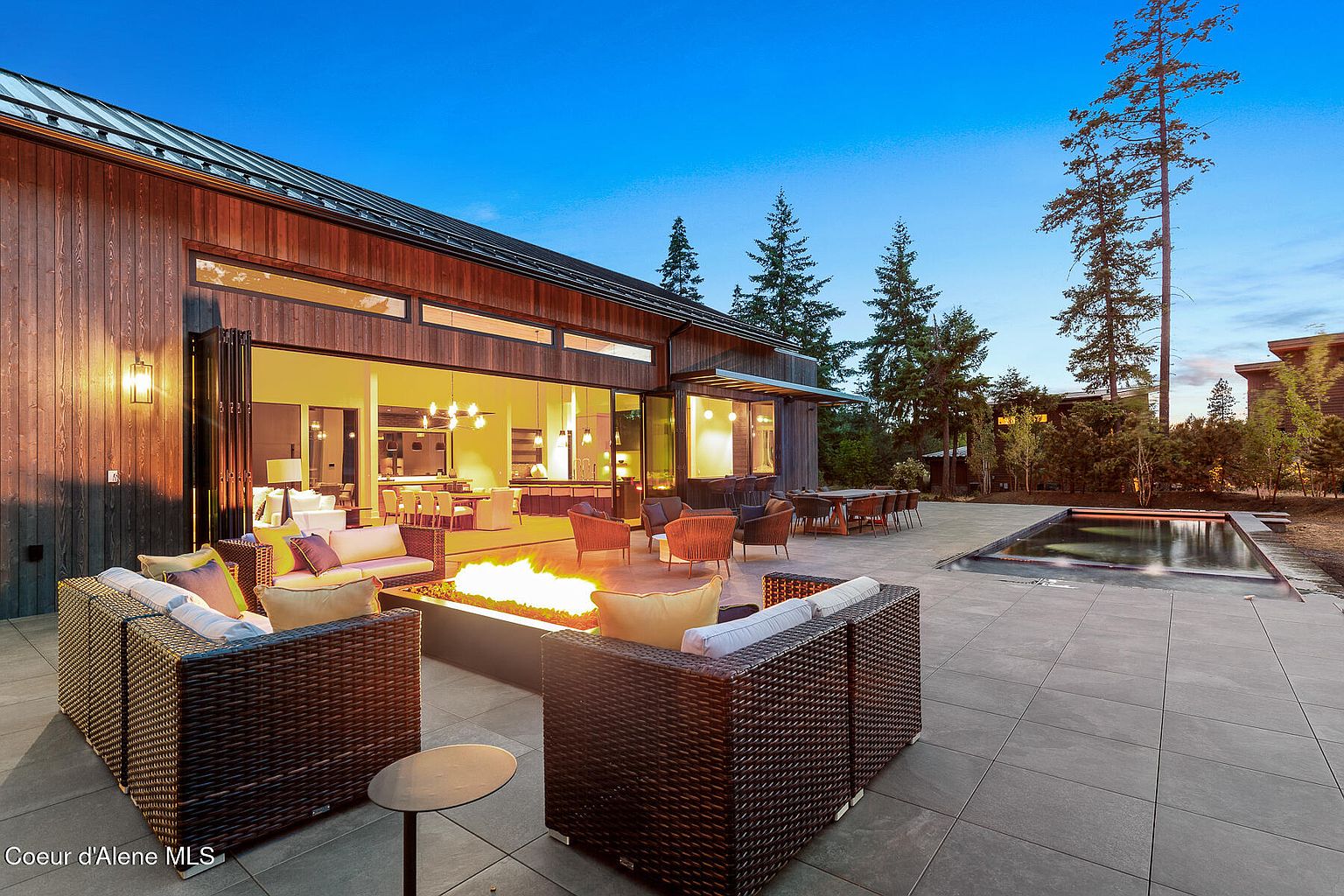This image showcases a luxurious outdoor patio area featuring modern wicker furniture arranged around a fire pit. The patio seamlessly connects to the interior through large, folding glass doors, creating an open and inviting space. A sleek pool is visible in the background, surrounded by well-maintained landscaping, enhancing the property's appeal for outdoor living and entertaining.