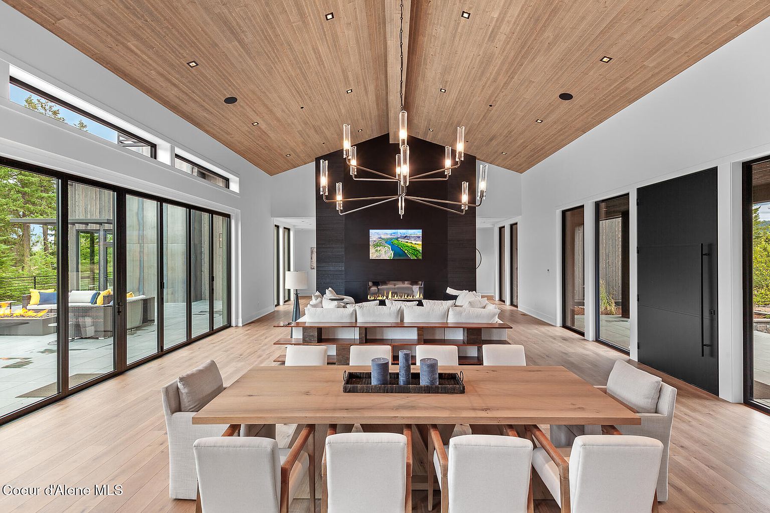 This is an interior shot of a dining room that flows into a living room. The dining area features a large wooden table with upholstered chairs, while the living room has a modern fireplace and comfortable seating. The space is well-lit with natural light from large windows and a modern chandelier, creating an open and inviting atmosphere.