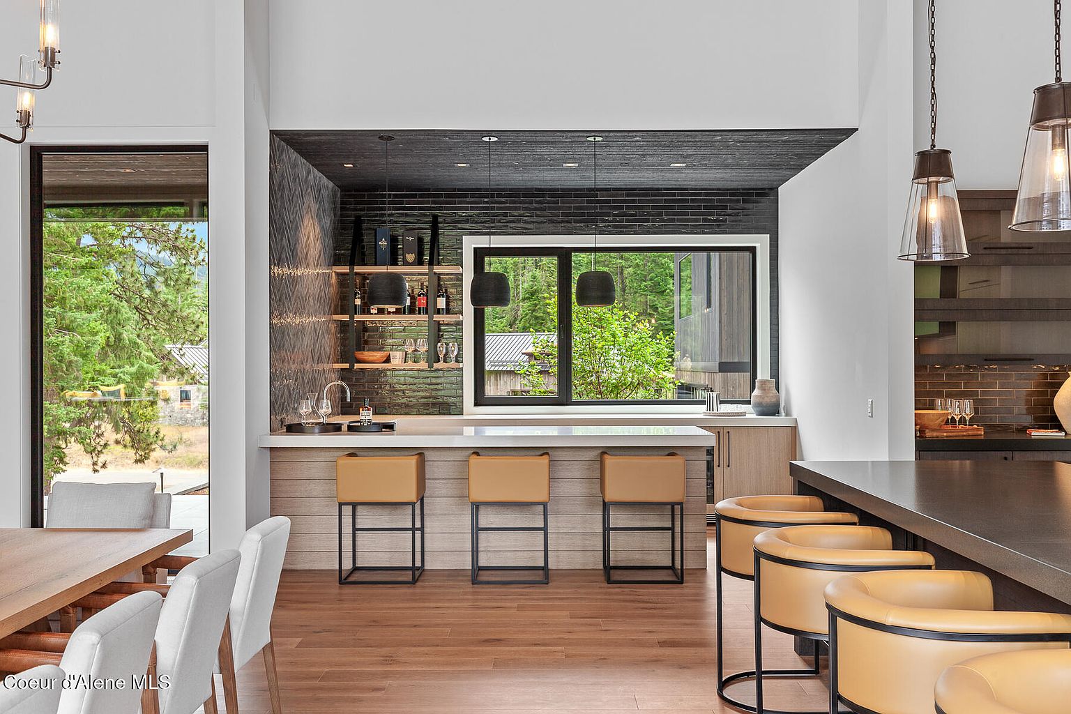 This interior shot showcases a modern kitchen with a stylish bar area. The bar features a light wood finish, complemented by tan leather bar stools with black frames. A window above the bar provides natural light and a view of the outdoors, while pendant lights illuminate the space, creating a warm and inviting atmosphere.