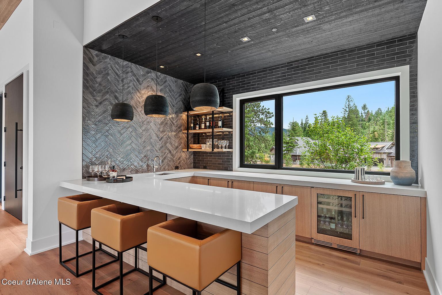 This interior shot showcases a stylish wine cellar or bar area, featuring a white countertop with seating, modern pendant lighting, and a built-in wine refrigerator. The backsplash and ceiling are adorned with dark, textured materials, adding a sophisticated touch. A window provides natural light and a view of the outdoors, enhancing the ambiance.