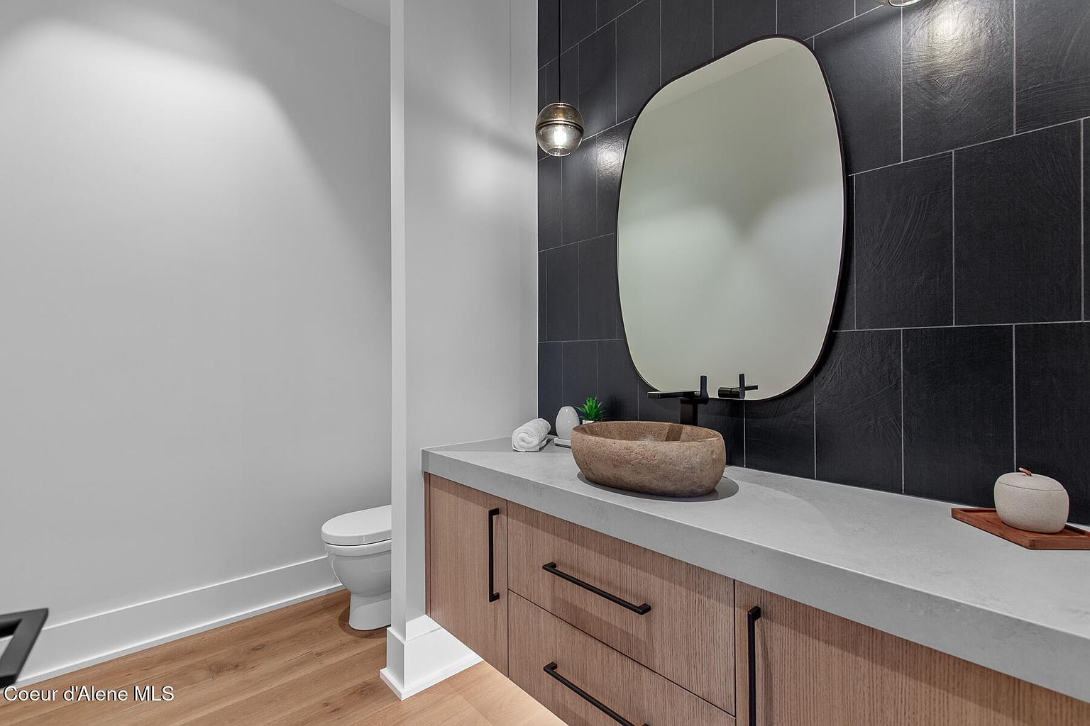 This is a modern guest bathroom featuring a unique stone vessel sink atop a light wood vanity with sleek, dark hardware. A large, organically shaped mirror hangs against a dark tiled wall, complemented by a minimalist pendant light. The space is clean and contemporary, with a toilet visible to the side and light wood flooring.
