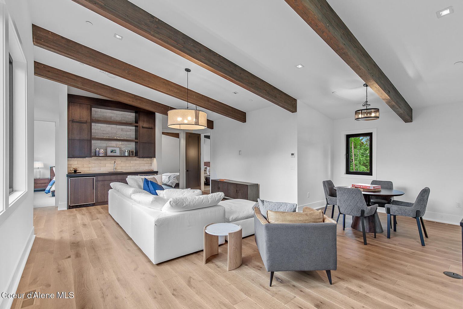 This is an interior shot of a living room featuring hardwood floors, exposed wooden beams, and white walls. The room includes a white sectional sofa, a gray armchair, a round dining table with gray chairs, and a built-in bar area with dark wood cabinetry. The overall impression is modern and inviting, with a focus on natural light and comfortable living spaces.