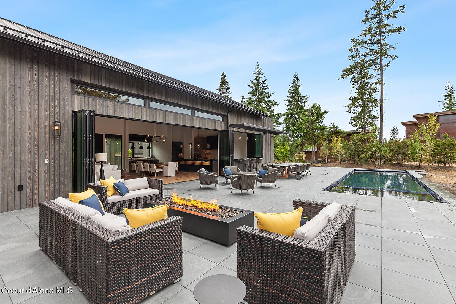 This image showcases a luxurious outdoor patio area, complete with comfortable wicker seating arranged around a modern fire pit. The patio seamlessly connects to the home through large, retractable glass doors, offering an open-concept feel. A swimming pool is visible in the background, enhancing the property's appeal for outdoor living and entertaining.