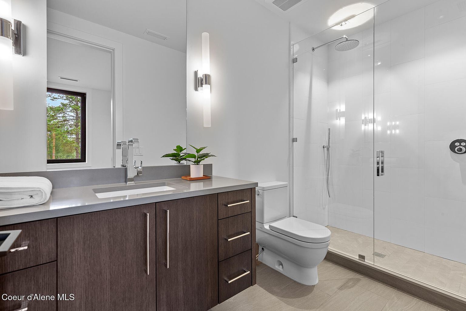 This is a modern bathroom featuring a dark wood vanity with a gray countertop, a rectangular sink, and a sleek faucet. A large mirror reflects a window, and modern light fixtures illuminate the space. The bathroom also includes a toilet and a glass-enclosed shower with white tile walls and a tiled floor, creating a clean and contemporary aesthetic.