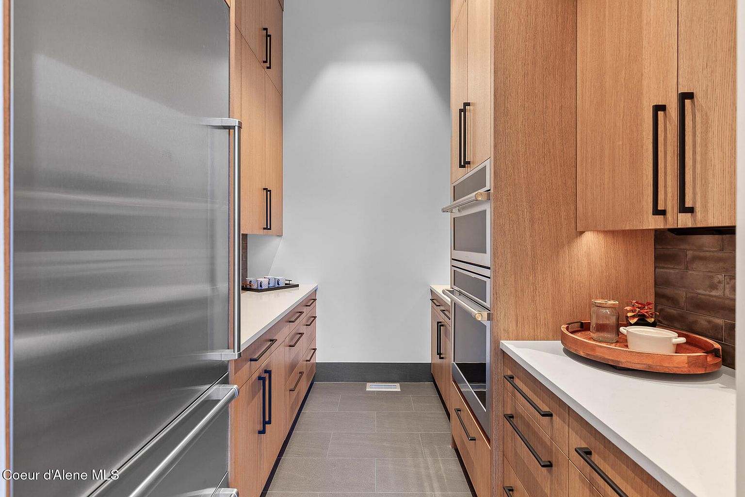 This is a well-organized pantry featuring light wood cabinetry with sleek, dark hardware. Stainless steel appliances, including a refrigerator and built-in ovens, add a modern touch. The countertops are light-colored, and the floor is tiled in a neutral tone, creating a clean and functional space.