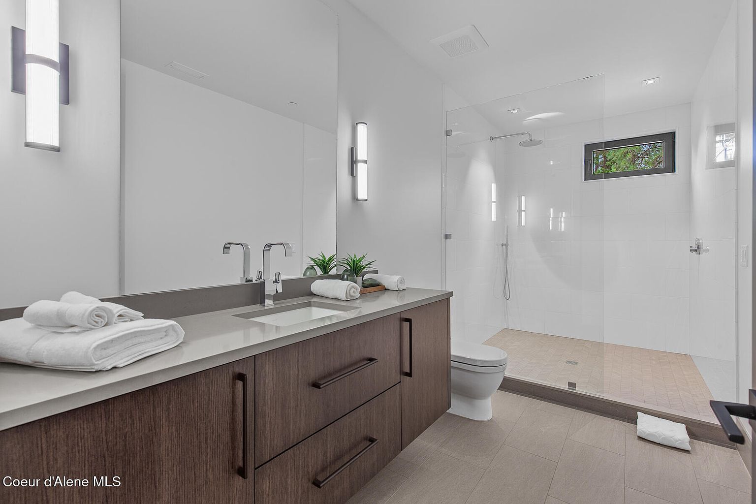 This is a modern primary bathroom featuring a double vanity with a gray countertop and dark wood cabinets. The bathroom includes a glass-enclosed shower with white tile and a small window. The overall design is clean and contemporary, with a neutral color palette and minimalist fixtures.