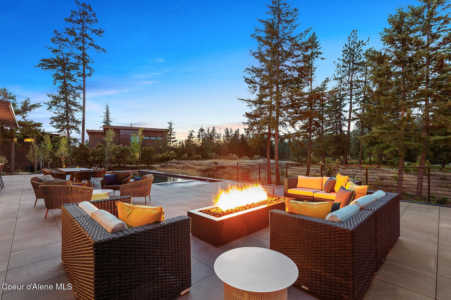 This image showcases a luxurious outdoor patio area, perfect for entertaining or relaxing. The space features comfortable wicker seating arranged around a modern fire pit, creating a cozy ambiance. A swimming pool is visible in the background, along with mature trees and a contemporary home design, enhancing the property's appeal.