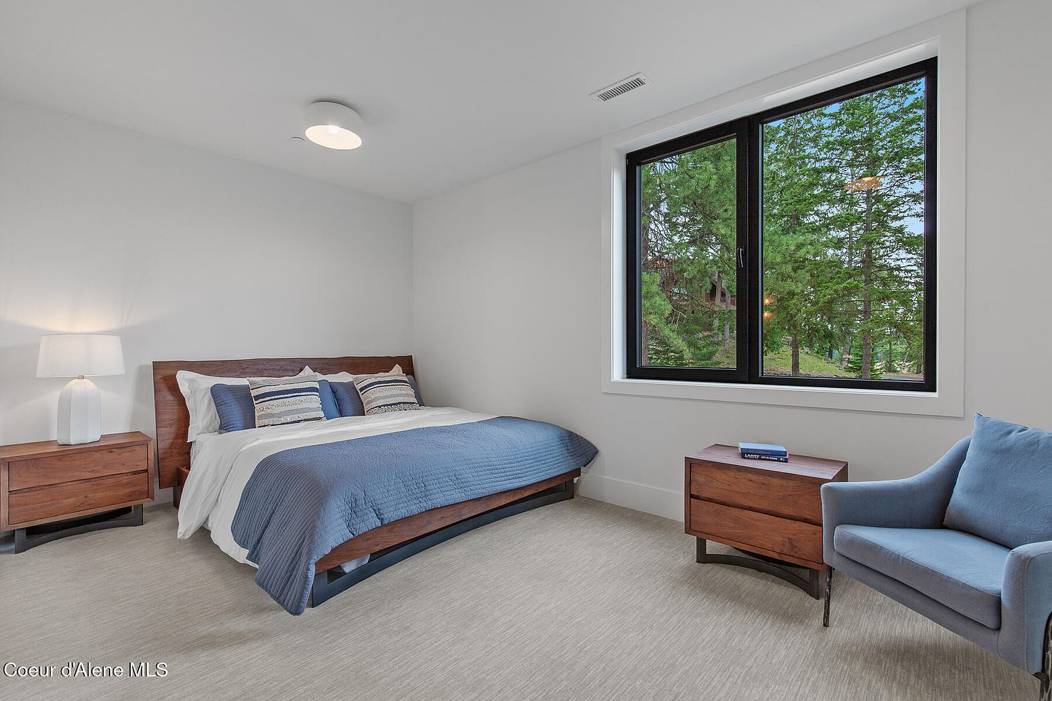 This is a bright and airy guest bedroom featuring a wooden bed frame with blue bedding, complemented by a matching blue armchair. A large window with a black frame offers a view of the surrounding trees, enhancing the room's tranquil atmosphere. The room is simply furnished with wooden nightstands and a modern ceiling light, creating a clean and inviting space.