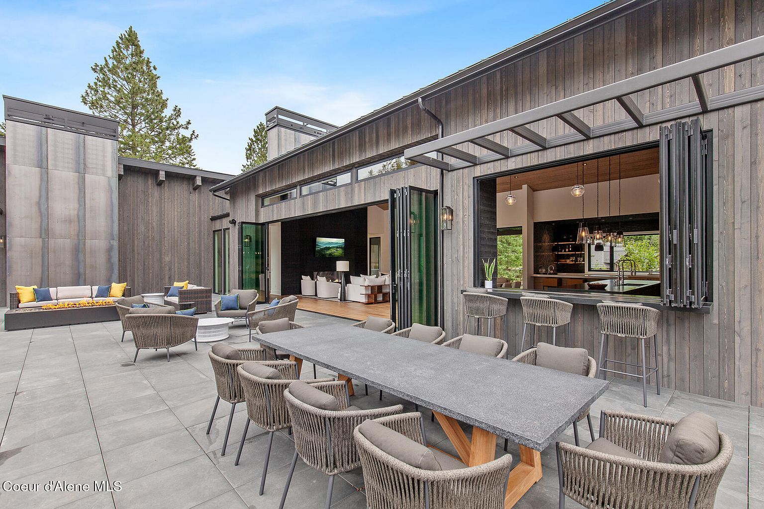 This image showcases a modern patio area, featuring a large outdoor dining table with seating for ten, a comfortable lounge area with a fire pit, and an open-air bar area connected to the interior kitchen. The patio is paved with large gray tiles, and the exterior of the house is clad in dark wood, creating a sophisticated and inviting outdoor living space.