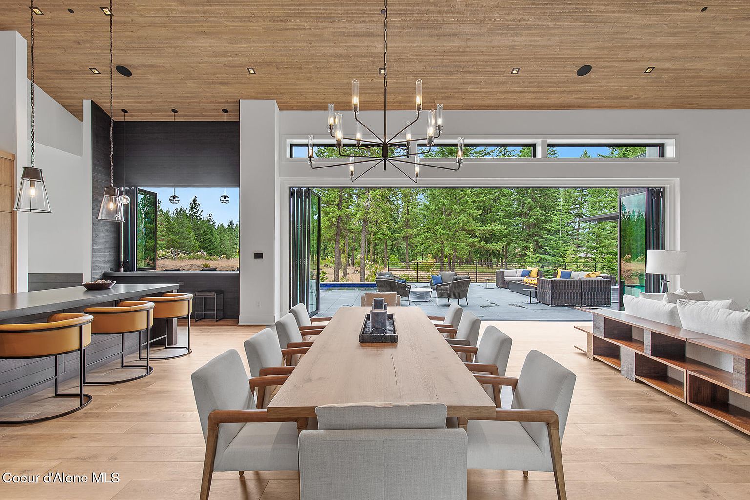 This is a bright and airy dining room featuring a long wooden table with modern chairs. The room opens to an outdoor patio through large folding glass doors, offering seamless indoor-outdoor living. A contemporary chandelier hangs above the table, and the space is filled with natural light, creating an inviting atmosphere.