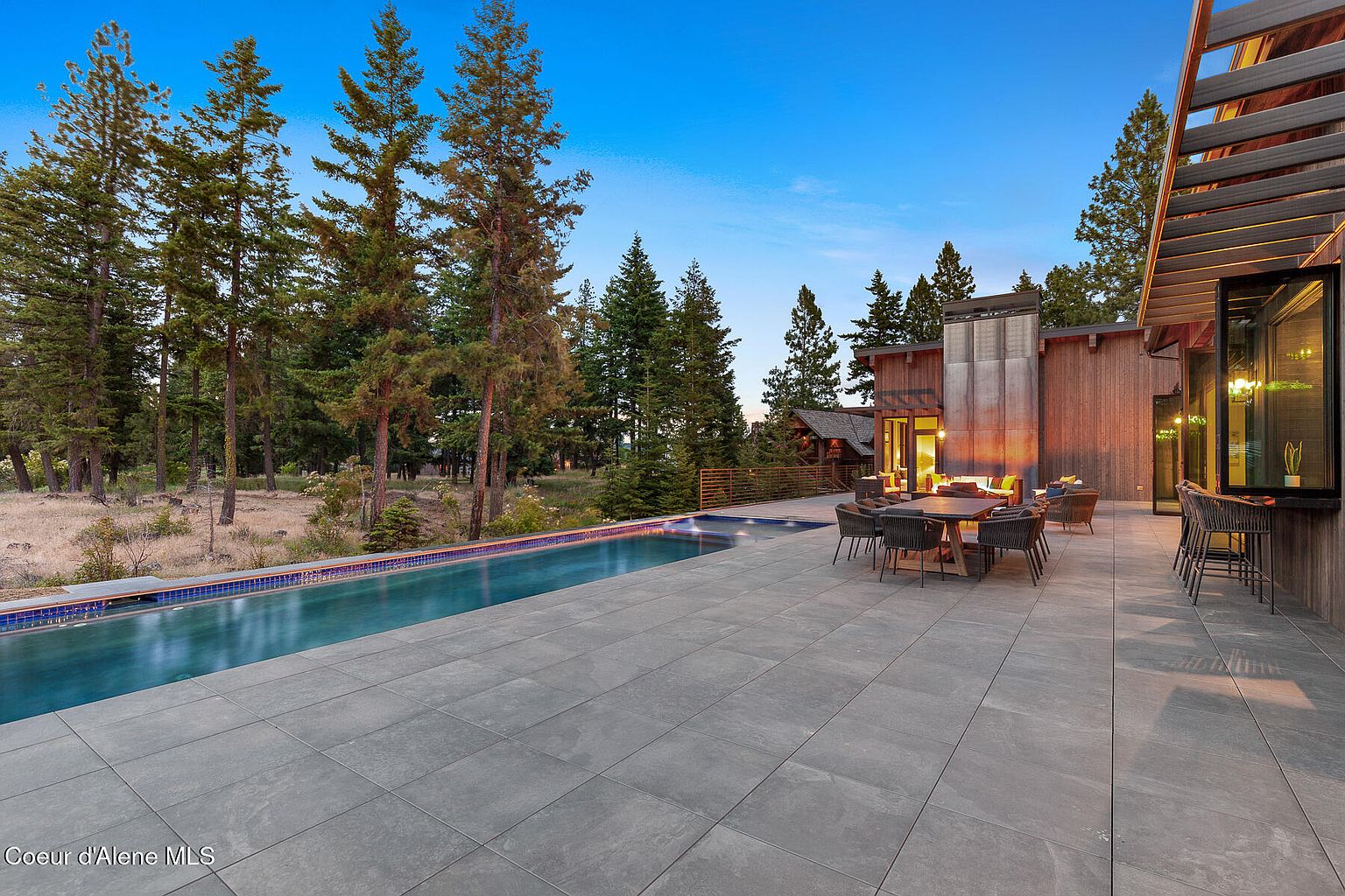This image showcases a luxurious outdoor space featuring a long, rectangular pool with a tiled edge, surrounded by a spacious gray tiled patio. Adjacent to the pool is an outdoor dining area with a large table and chairs, as well as a lounge area with comfortable seating. The backdrop includes mature trees and a modern home with wood siding and architectural details, creating a serene and upscale atmosphere.