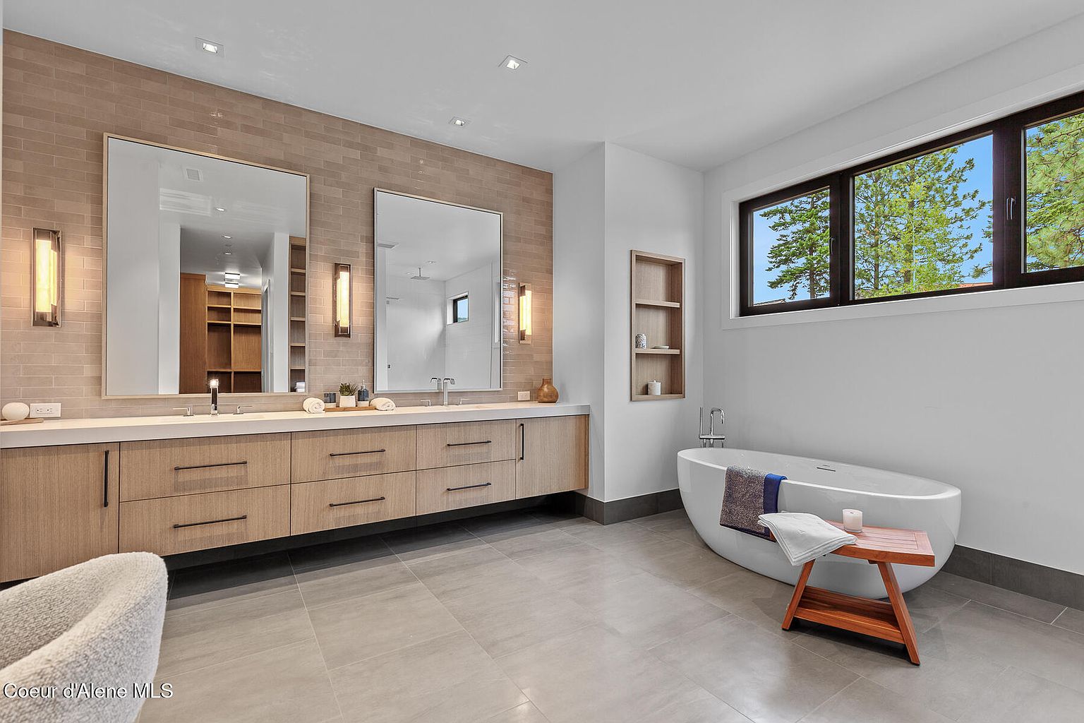 This is a luxurious primary bathroom featuring a double vanity with light wood cabinetry and a white countertop, complemented by a brick-style tiled wall. Large mirrors are flanked by sconce lighting, and a freestanding bathtub sits beneath a window offering natural light and a view of trees. The overall impression is modern, clean, and spa-like.