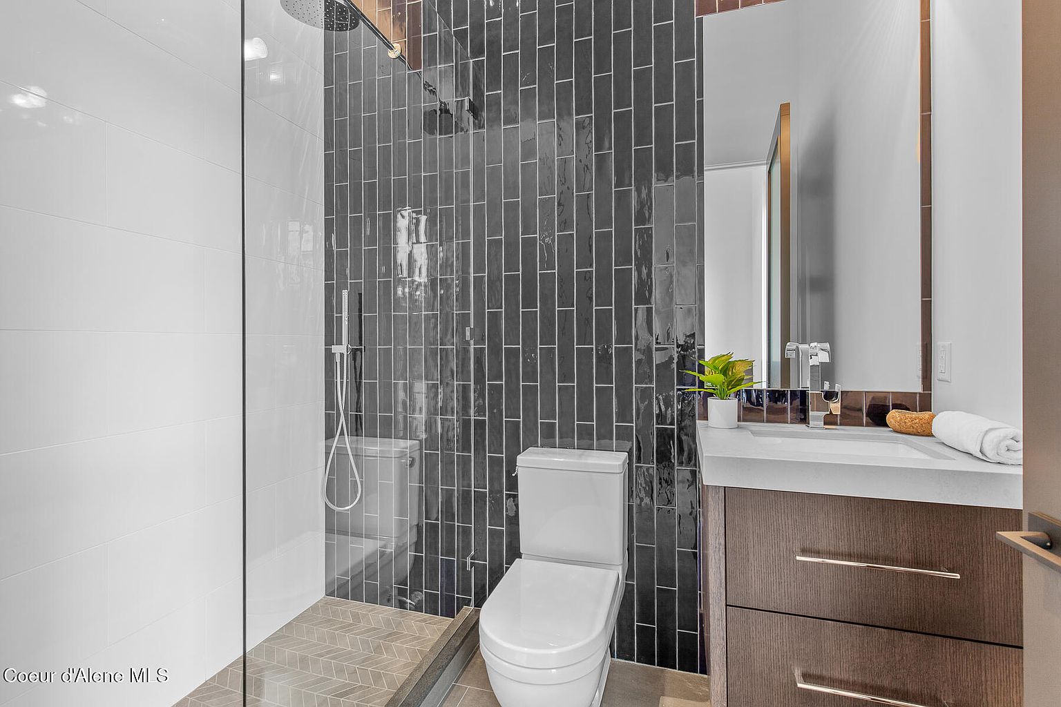 This is a modern bathroom featuring a walk-in shower with white and dark gray vertical tiles, a toilet, and a vanity with a white countertop and dark wood cabinets. The shower has a glass enclosure and a rainfall showerhead. The overall style is contemporary and sleek, creating a clean and inviting space.